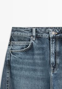 Denim jeans tailleband met riemlus, metalen knoop, voorkant linkerpocket en zichtbare stiksels op verweerd blauw stof.