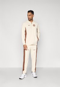 Crème zip-up hoodie met bruine accenten en logo, gecombineerd met crème joggers met bruine zijkanten en merklogo, voltooid met witte sneakers.