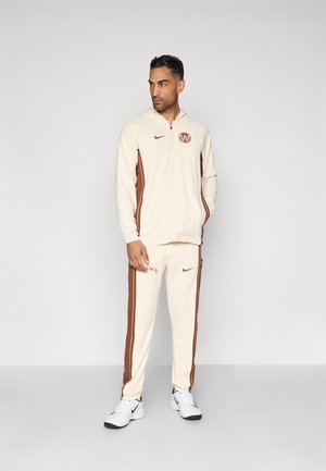 Krem fôrer med brune detaljer og logo, parret med krem joggebukser med brune sidestriper og merke-logo, fullført med hvite sneakers.