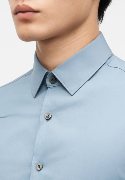 Κοντινό πλάνο ενός ατόμου που φορά ένα γαλάζιο πουκάμισο με γιακά, κουμπωμένο μέχρι το επάνω μέρος, δείχνοντας τον λαιμό, το πηγούνι και μέρος της γνάθου.