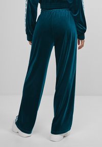 Kalhoty z veluru v tyrkysové barvě s volným střihem, vybavené elastickým pasem a kapsami na bočních švech. Ozdobené potiskem loga po straně.