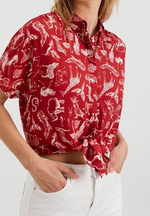 Rood overhemd met korte mouwen en witte diertjesillustraties, gestrikt in de taille, gedragen met een witte broek. Handen in de zakken.