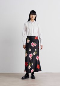 Svart blomstermønstret skjørt med rosa og røde blomster, sammen med en hvit skjorte med krage. Modellen har på seg svarte klumpete sko, stående på et lyst gulv.