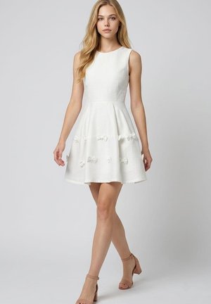 Mujer joven caminando, vestida con un vestido blanco sin mangas con pequeños lazos y tacones beiges abiertos en la punta con correa en el tobillo, sobre un fondo gris liso.