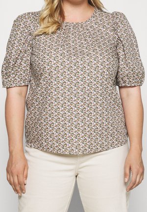 Femme portant un chemisier à manches courtes à motif floral avec des manches bouffantes et un pantalon beige clair, debout devant un fond uni.