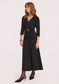 Czarna sukienka z długim rękawem, dekoltem w kształcie litery V, z marszczonym przodem i szczegółowym złotym paskiem. Noszona z przezroczystymi rajstopami i szpilkami w szpic.