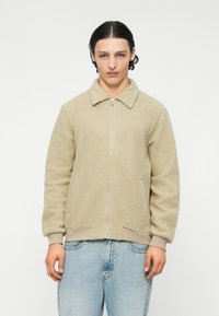 Giovane con capelli scuri tirati indietro, indossa una giacca a zip beige in pile e jeans azzurri chiari, in piedi contro uno sfondo semplice.