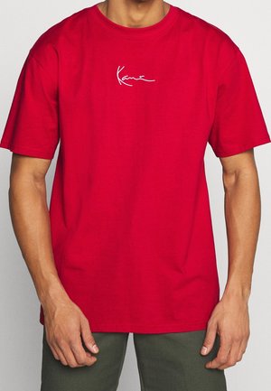 Camiseta roja de algodón con mangas cortas y un corte holgado. Presenta un logo blanco de firma en el pecho. Textura suave, sin patrones.