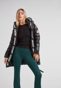 Mujer vistiendo un abrigo de plumas negro con la letra "JACKIT" en el interior, top sin mangas negro y pantalones de campana teal, de pie contra un fondo simple.