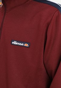 Bordeauxkleurige zip-up jack met zwart-wit Ellesse logo patch op de borst en een zwarte streep op de schouder.