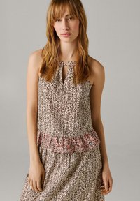 Jonge vrouw met steil lichtbruin haar, gekleed in een mouwloze jurk met patroon en een gerimpeld detail ter hoogte van de taille, staand tegen een effen achtergrond.