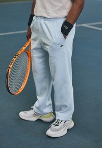 Világoskék melegítőnadrág, fekete Lacoste logóval, szürke és zöld sportcipőkkel, narancssárga teniszütőt tartva egy kék pályán.