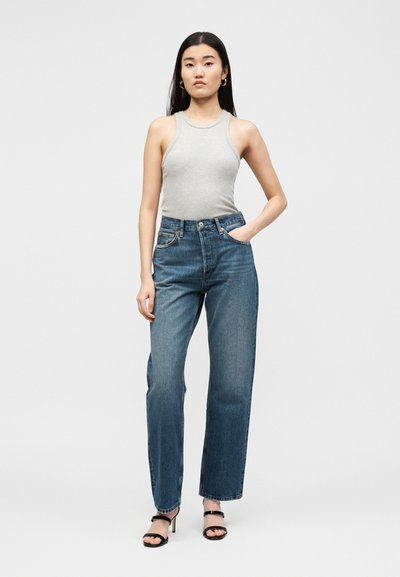 Νέα γυναίκα που στέκεται φορώντας γκρι αμάνικο ριμπ τοπ, τζιν με ψηλή μέση σε μπλε χρώμα, μαύρα ανοιχτά τακούνια, με το ένα χέρι στην τσέπη, μπροστά από απλό φόντο.
