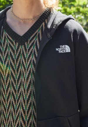 Osoba nosząca sweter z wzorem w zygzaki w kolorach zielonym, beżowym i czarnym pod czarną rozpinaną bluzą North Face na zewnątrz.