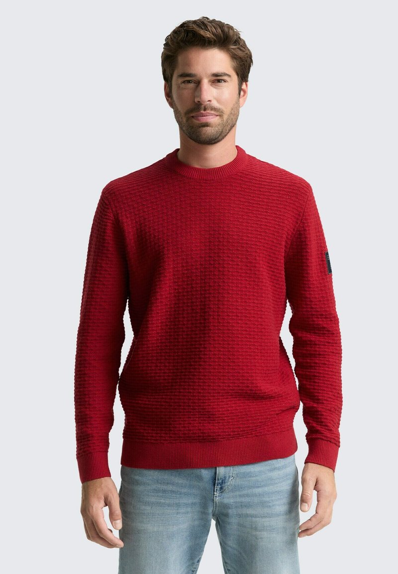 Rode textuurtrui met ronde hals en geribbelde zoom. Bevat een subtiel patroon en logo op de mouw. Gepaarde met blauwe jeans.