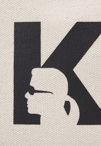 Czarna litera K z sylwetką kobiety noszącej okulary przeciwsłoneczne i kucyk na tle teksturowanej beżowej tkaniny.