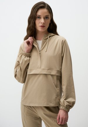 Jonge vrouw met lang golvend haar, gekleed in een beige hoodie met rits en bijpassende broek, die met één hand de kraag van de hoodie vasthoudt.