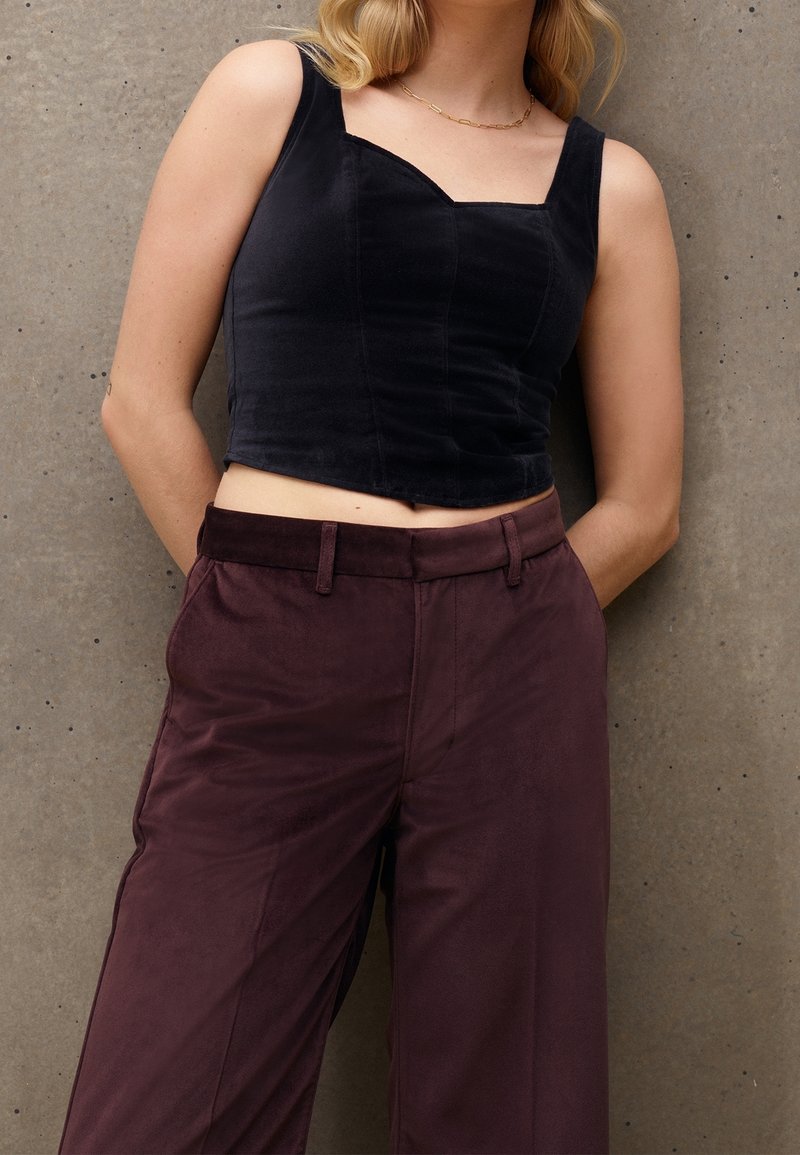 Femme portant un haut court sans manches noir et un pantalon taille haute violet foncé, debout devant un mur en béton gris.