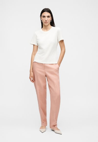 Γυναίκα στέκεται με το ένα χέρι στην τσέπη, φοράει λευκό κοντομάνικο πουκάμισο, ανοιχτό ροζ παντελόνι, λευκά μυτερά παπούτσια και ασημένια σκουλαρίκια κρίκους.