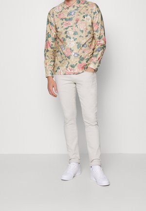 Homme portant une chemise à fleurs beige à manches longues, un pantalon slim crème et des baskets blanches, debout devant un fond uni clair.