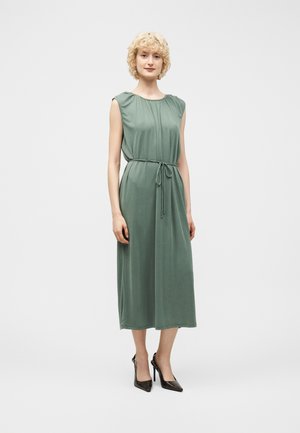 Kobieta o krótkich, kręconych blond włosach, ubrana w bezrękawową, szałwiowozieloną sukienkę midi z wiązaniem w talii oraz czarne szpilki, stojąca.