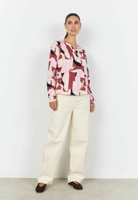 Lyserød bluse med lange ærmer og abstrakte mønstre i bordeaux, sort og creme; kombineret med cremefarvede bukser med vide ben og mørke sko.