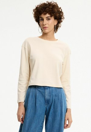Mujer con cabello rizado que lleva pendientes de aro, una blusa crema de manga larga y vaqueros azules sueltos de talle alto, de pie sobre un fondo blanco.