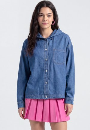 Junge Frau trägt eine blaue, knöpfbare Jeansjacke mit Kapuze und einen rosa plissierten Rock, steht vor einem einfarbigen Hintergrund.