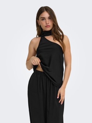 Femme aux longs cheveux bruns portant un top noir sans manches à col halter et un pantalon noir, tenant l'ourlet du top d'une main.