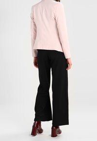 Ljusrosa figursydd kavaj med rund hals, parat med svarta vida byxor. Modellen bär bordeauxfärgade klackstövlar med tjock klack.