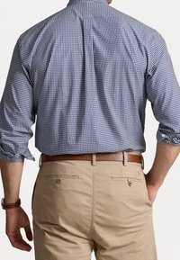 Mężczyzna w niebieskiej koszuli w kratę z podwiniętymi rękawami, wciągniętej w beżowe spodnie, z brązowym skórzanym paskiem i zegarkiem na ręce, z ręką w kieszeni.