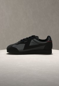 Μαύρο αθλητικό παπούτσι χαμηλής κοπής με υφασμάτινα πάνελ, κορδόνια στο μπροστινό μέρος και διακριτική κόκκινη λεπτομέρεια στη φτέρνα, εμφανιζόμενο από την πλευρά σε ουδέτερη επιφάνεια.