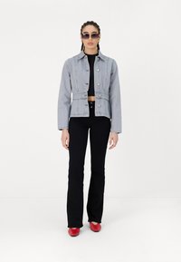 Veste en denim cropped bleu clair avec des boutons sur le devant, associée à un pantalon noir évasé et des chaussures rouges. Les accessoires comprennent des lunettes de soleil oversize.