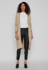 Cardigan en tricot beige avec ouverture à l'avant et poignets côtelés, porté sur une chemise blanche, associé à un pantalon noir slim-fit et des mules noires à talons.