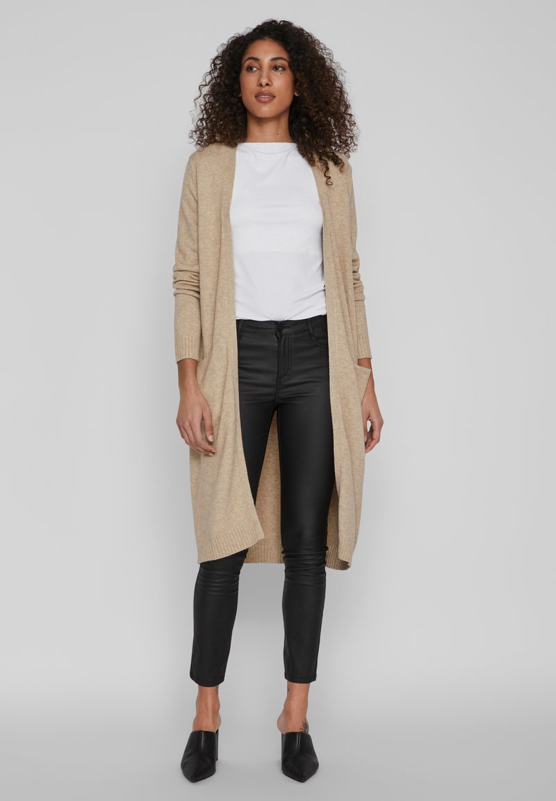 Cardigan en tricot beige avec ouverture à l'avant et poignets côtelés, porté sur une chemise blanche, associé à un pantalon noir slim-fit et des mules noires à talons.