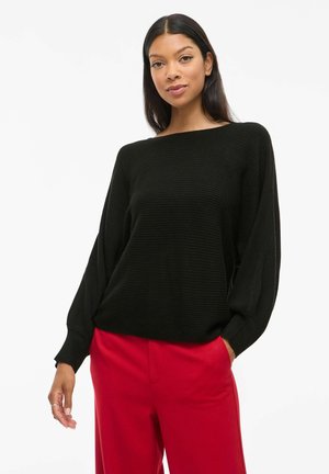 Pull en tricot noir avec texture côtelée, col bateau large et manches longues, associé à un pantalon rouge ajusté.