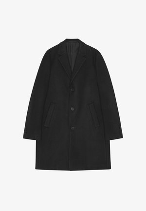 Svart ullrock med en skarpt skuren krage, tre knappar framtill, sidfickor och knappdetaljer på ärmarna.