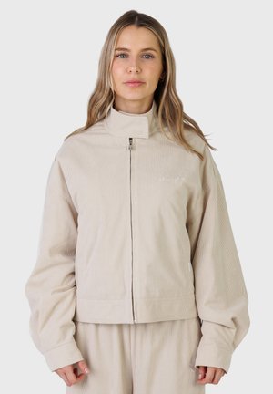 Giovane donna con lunghi capelli biondi che indossa una giacca beige in velluto a coste con zip e pantaloni abbinati, in piedi davanti a uno sfondo semplice.