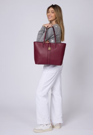 Bolso tote burdeos con dos asas, detalle de cierre dorado, textura suave y forma estructurada. Llevado sobre un suéter gris y pantalones blancos.