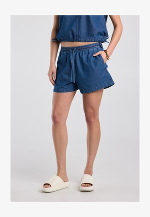 Vrouw draagt blauwe denim shorts met trekkoord en bijpassende mouwloze top met witte slip-on sandalen, staand met één hand in de zak.