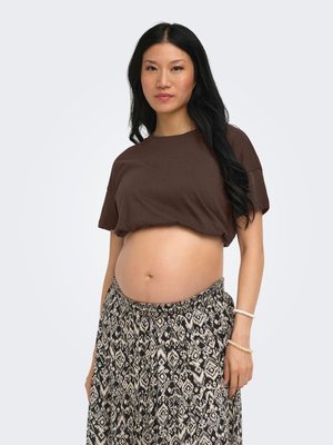 Jeune femme aux longs cheveux noirs portant un t-shirt court marron et une jupe à motifs, montrant son ventre et debout devant un fond uni.