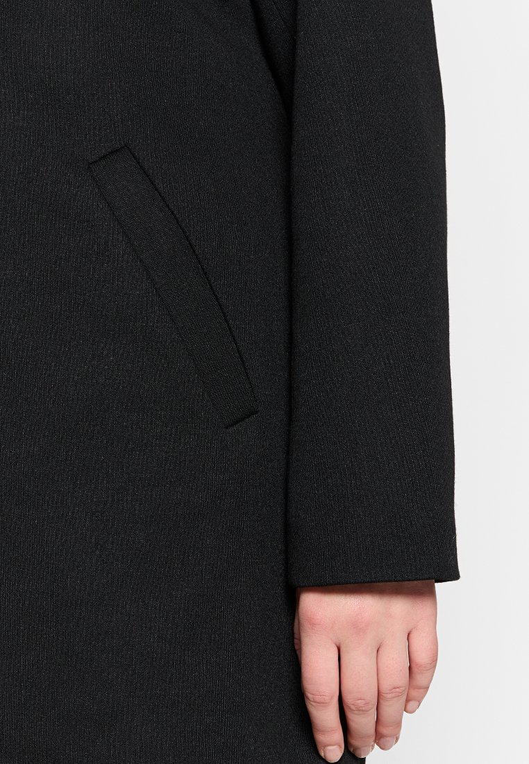 Cappotto nero in un tessuto texturizzato, con una tasca laterale e una manica dritta e pulita. Mano visibile nell'inquadratura.