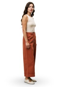 Pantalons en coton de couleur rouille, à jambes larges, dotés d'une taille haute et d'un ourlet net. Associés à un débardeur beige à côtes et à des chaussures claires.