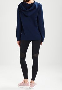 Marineblaue Kapuzenjacke mit Kapuze, kombiniert mit schwarzen Leggings mit Netzpaneelen. Dazu werden hellrosa, strukturierten Sneakers getragen.