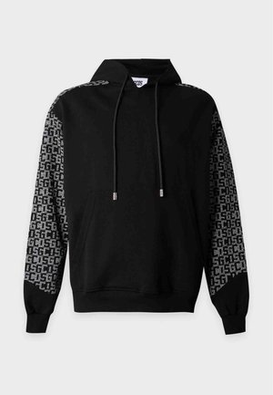 Jeune homme debout portant un sweat à capuche noir avec des manches et un ourlet à motifs, un pantalon noir et des baskets blanches sur un fond blanc uni.