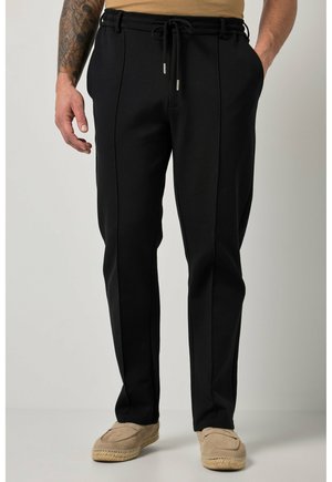 Homme portant un pantalon noir à cordon avec des coutures avant, des chaussures beiges à enfiler, et un bras tatoué visible sur un fond uni.