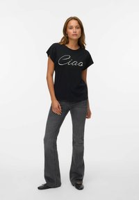 T-shirt noir à manches courtes avec un design en strass "Ciao", associé à un jean gris évasé et des chaussures noires sur fond blanc.