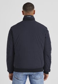 Marineblauer Bomberjacke mit hohem Kragen, elastischen Bündchen und Taillenbund, glatter Textur und Logo-Detail am Hals.