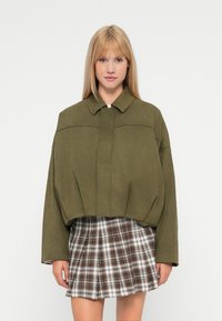 Giacca cropped verde oliva con colletto a punta, dal design ampio e chiusura frontale; abbinata a una mini gonna scozzese nei toni marrone e beige.