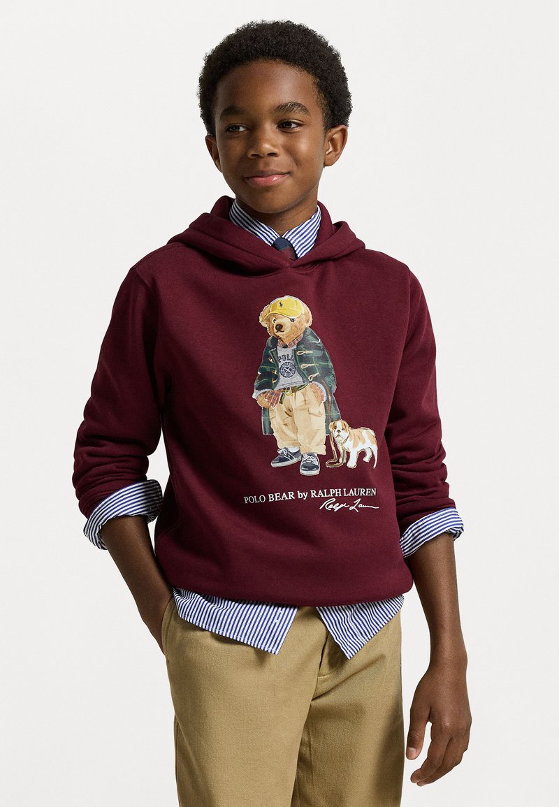 Bordo kapuca majica s grafikom medveda v zeleni jakni in svetlih hlačah, skupaj s psom. Obložena je z modro črtasto majico.